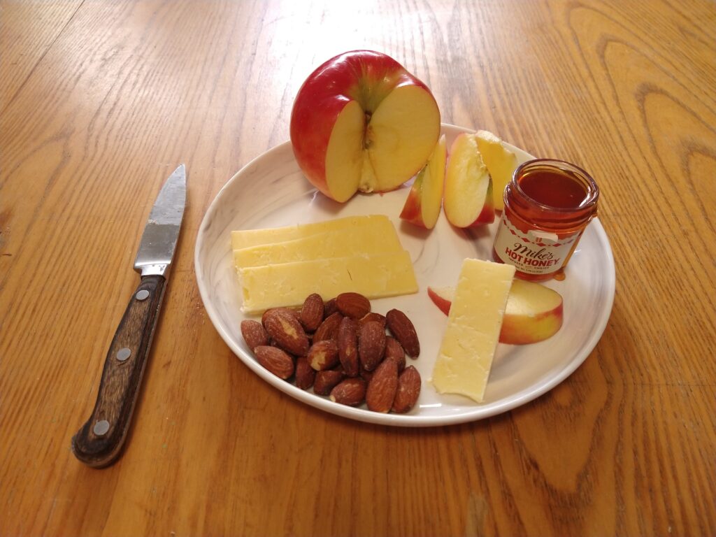 Plate containing a sectioned apple, slices of cheddar cheese, almonds, and a small pot of hot honey.